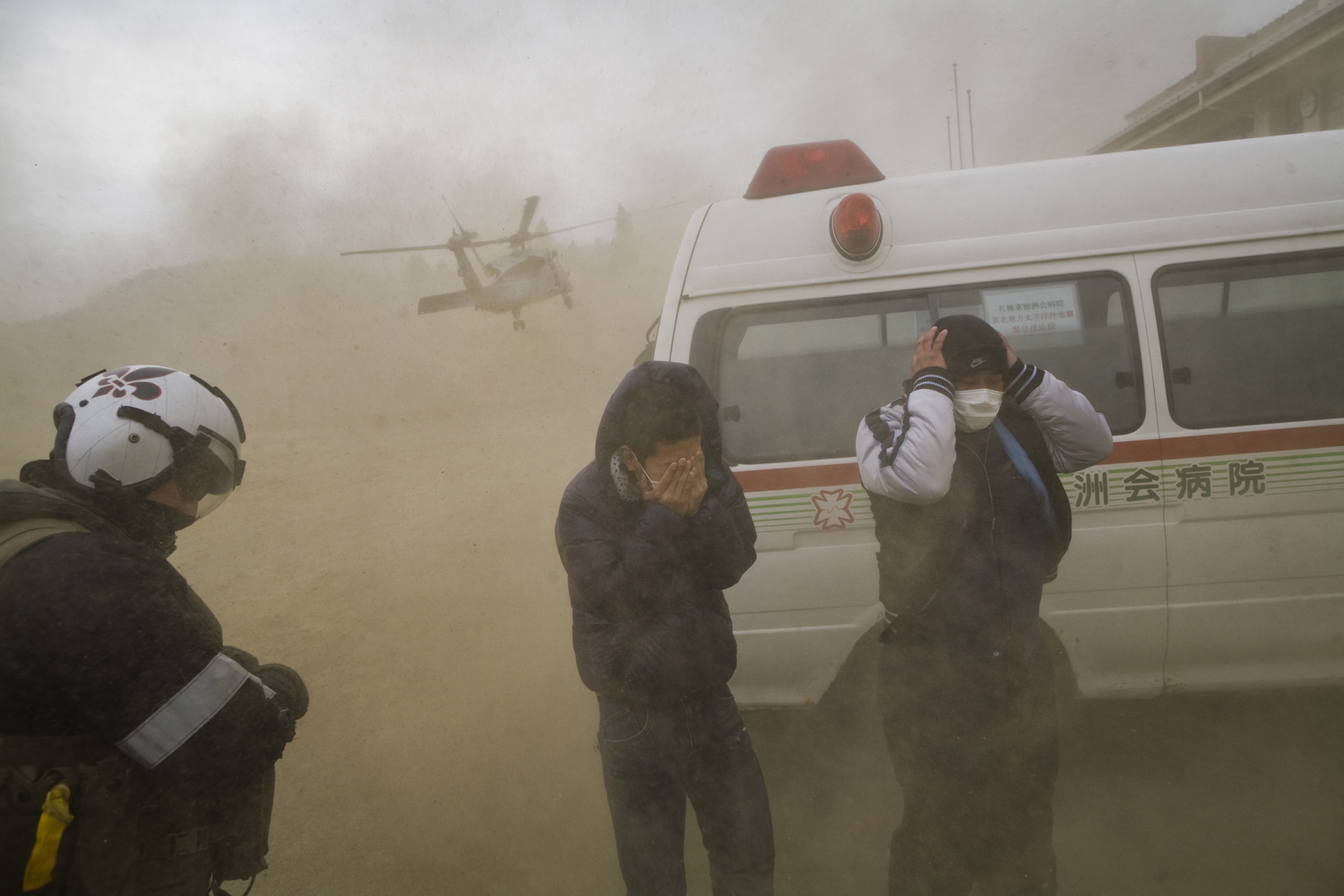 On the ground, workers protect themselves from dust as a helicopter with the Black Knights Helicopter Squadron delivering aid prepares to land in one of the damaged and isolated communities in the Sendai region. March 21, 2011