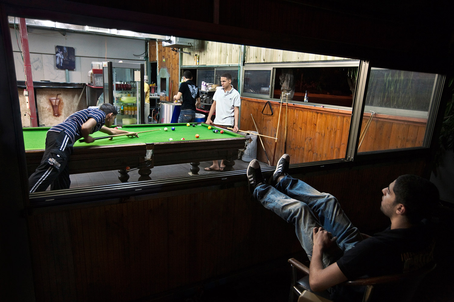 A young Arab man watches two of his friends playing pool in the local cafe in Jaljulia. Only 3% of the youth in Jaljulia complete their high school studies successfully and get their diploma. Most end up earning low wages working in construction for companies owned by Jews. The central city offers few recreation venues and most young men hang out in the streets or in the only cafe in town.