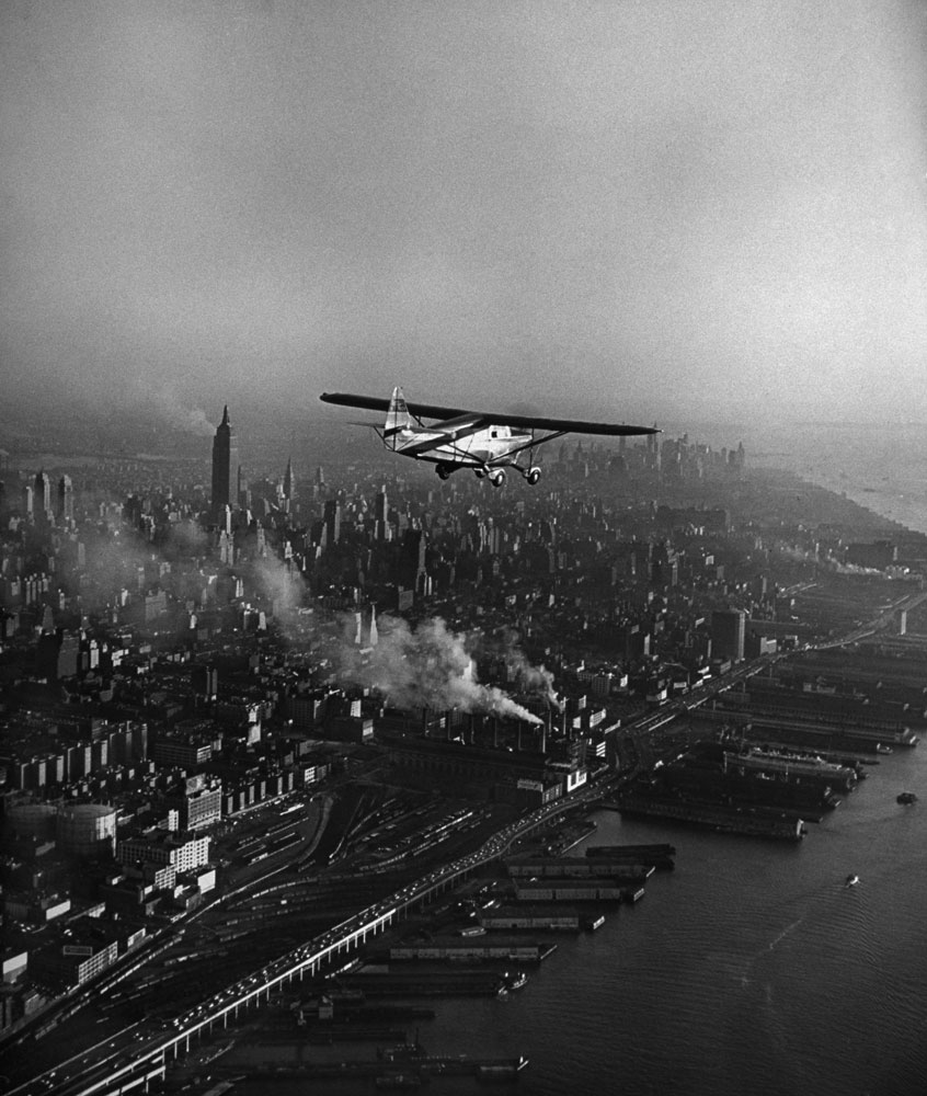 A plane flys above New York, NY before landing in October 1949.