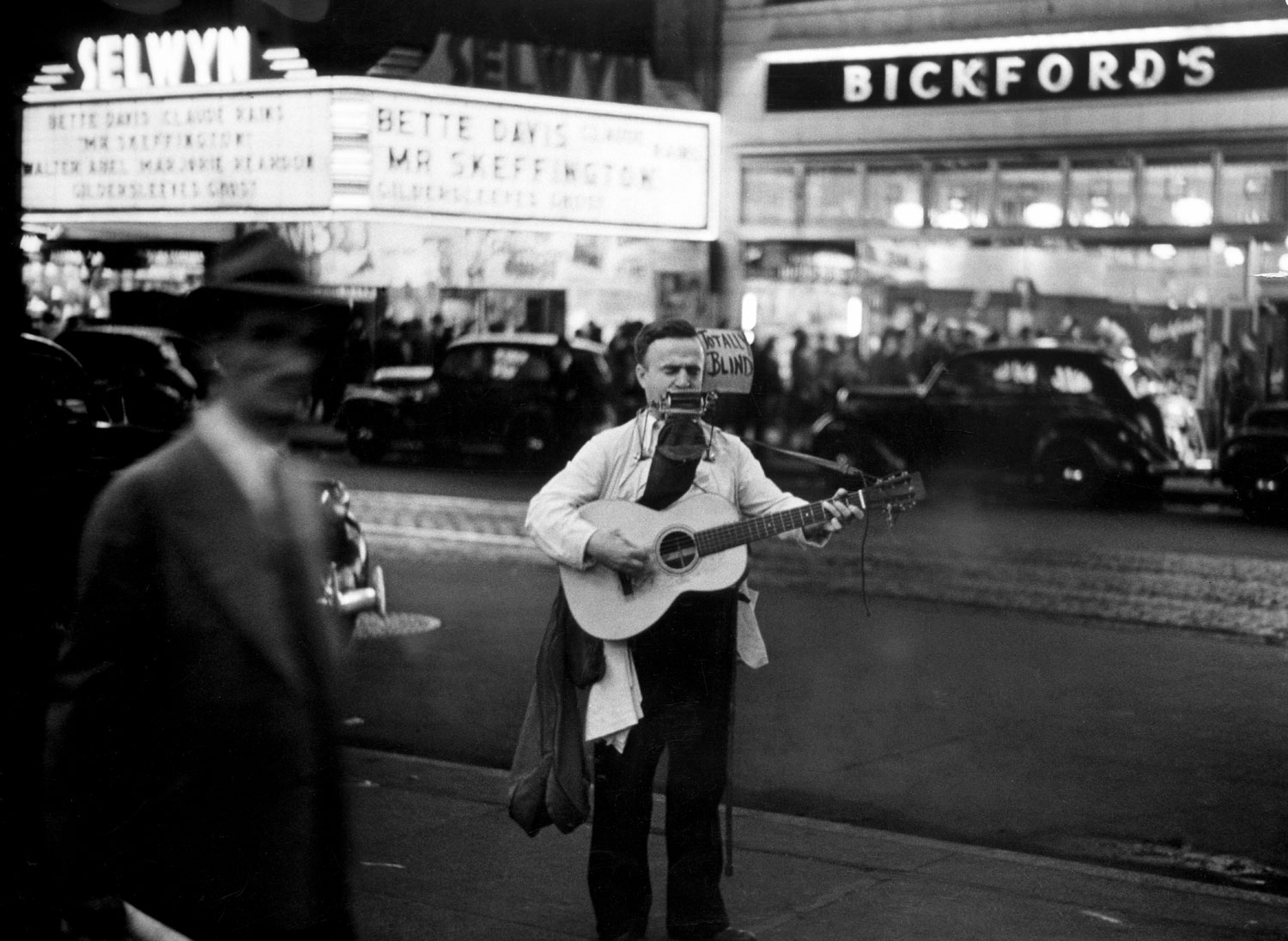 A strolling blind musician plays guitar and harmonica along Broadway at night in the Times Square Area in 1944. "Mr. Skeffington" is playing at the Selwyn Theater across the street.