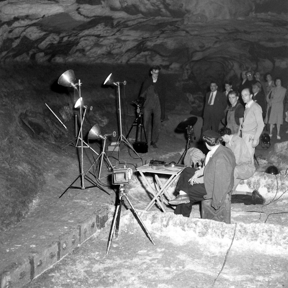 Ruth Morse sits at a desk where picture captions were written during the time that Ralph Morse made photographs at Lascaux, 1947. In the background, local townspeople see the cave paintings under bright lights for the very first time.