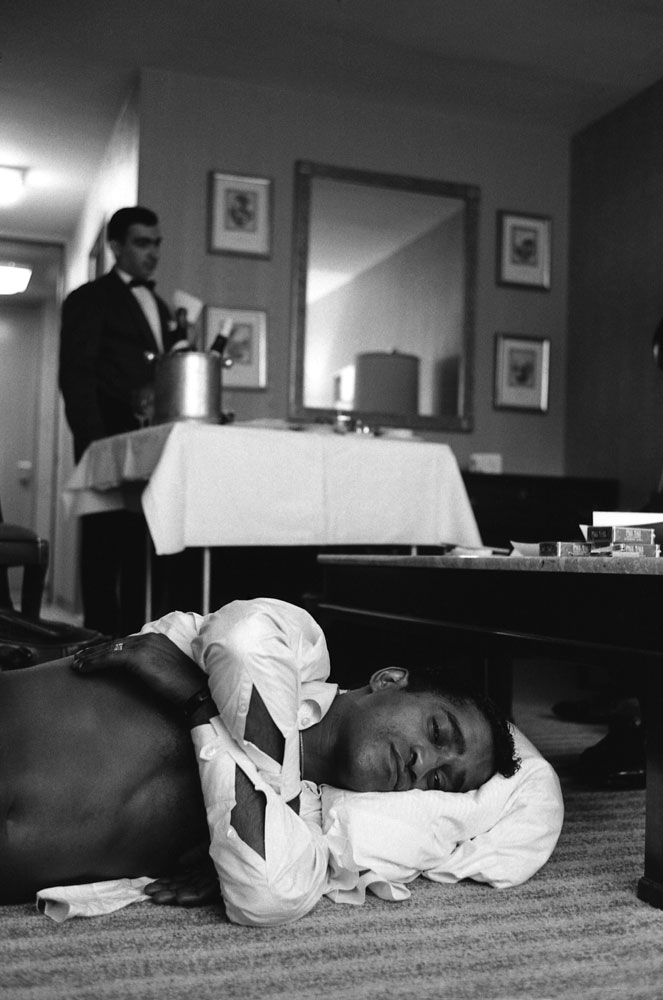 Sammy Davis Jr. rests on his side with a pillow on the floor of a New York City hotel room. A hotel staff member stands in the background with a room service cart.