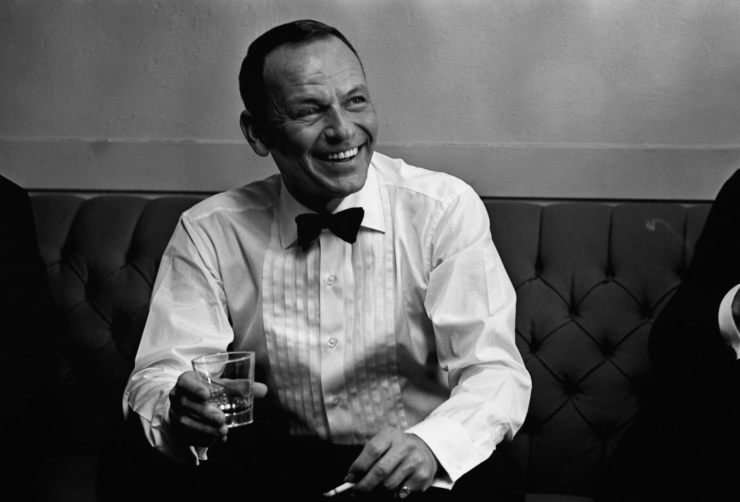 Portrait of Frank Sinatra in cigarette and high ball glass at the Sands Hotel and Casino in 1964. He is wearing a bow tie and tuxedo shirt and sitting on a sofa.