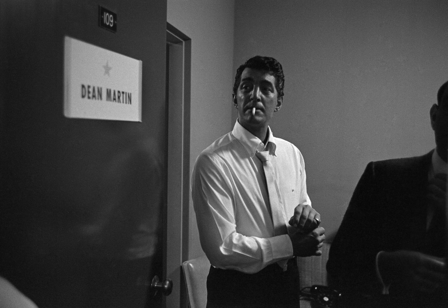 Dean Martin smokes a cigarette beside his dressing room door backstage before his performance in Las Vegas in 1958. He adjusts his cufflinks.
