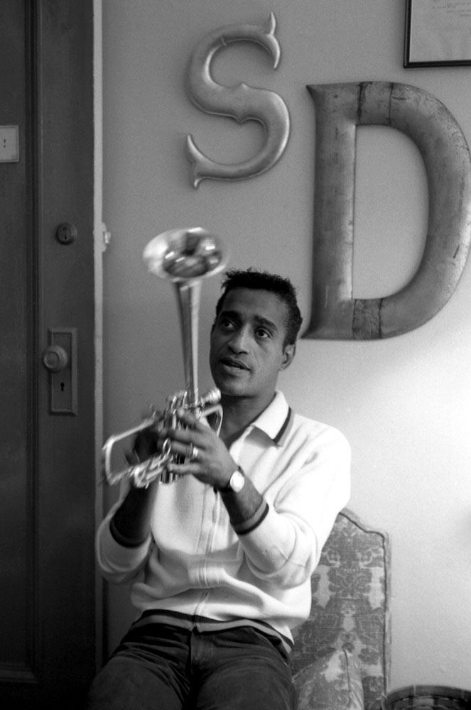 Sammy Davis Jr. plays with trumpet. The letters S and D hang on the wall behind him.