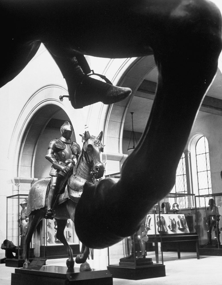Past the legs of a horse and rider is seen the Hall of Armor at the Metropolitan Museum of Art.