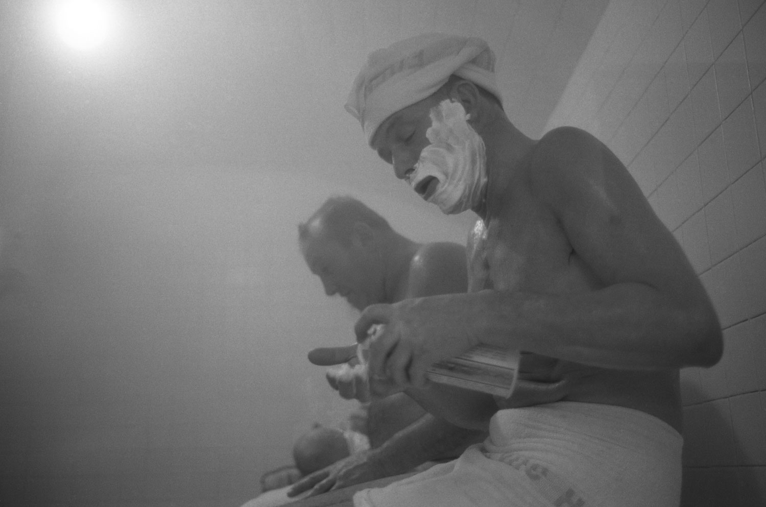 Frank Sinatra shaves in a steam room in Miami. He is wearing a towel around his waste and on his head. His face is covered with shaving cream.