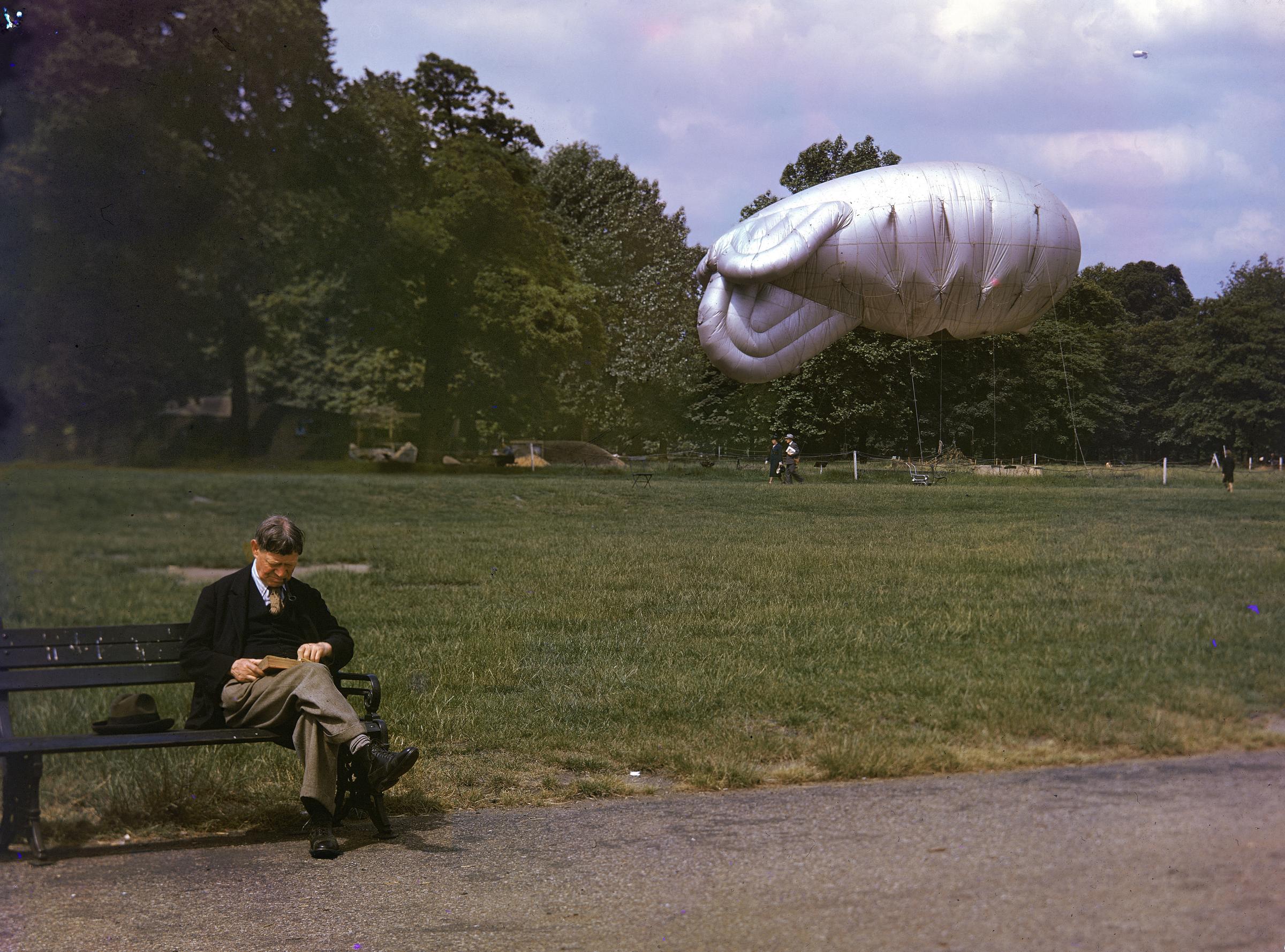 A man sits on a park bench in London, reading a book, 1940; a moored "barrage balloon" is visible in the background, while a second one soars high in the distance.
