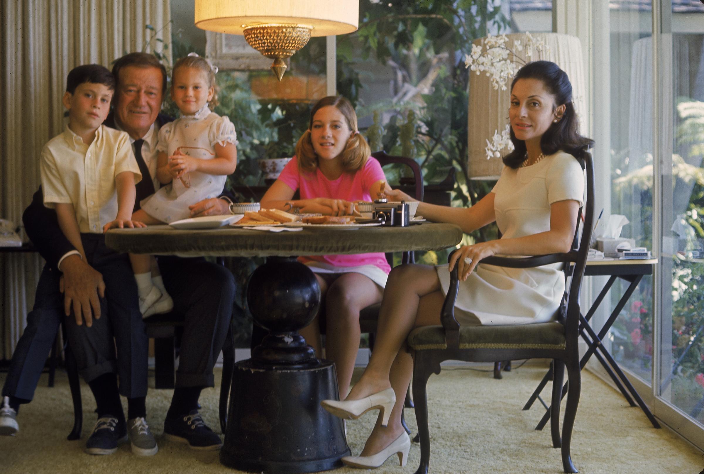 John Wayne with kids Ethan, Marissa, and Aissa and wife Pilar at dining table of family home in 1969