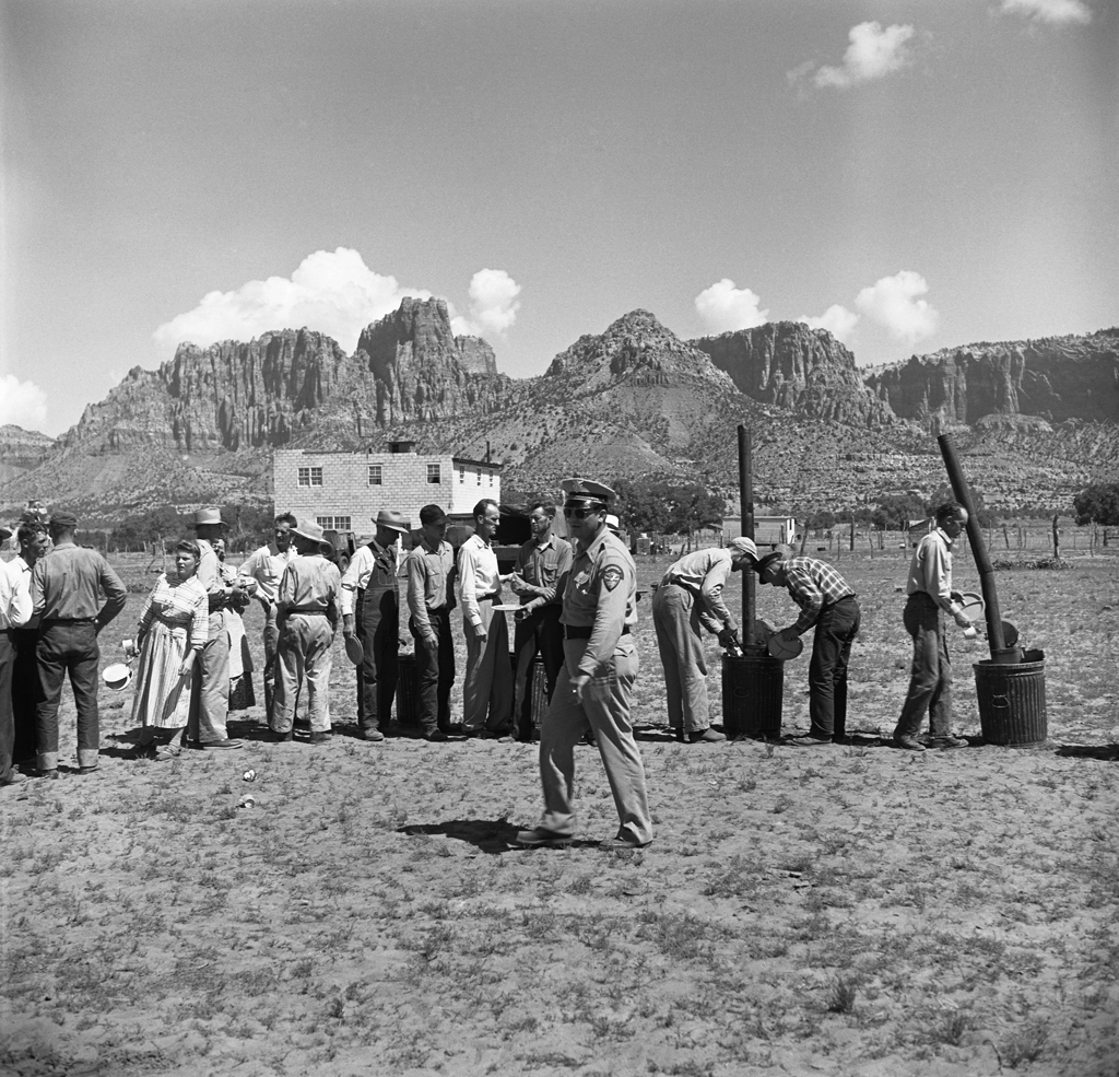 Under an officer's supervision, arrested polygamists line up beneath jagged Arizona cliffs, 1953.