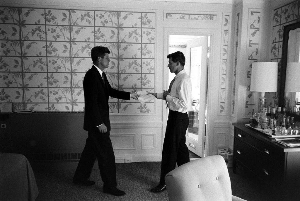 The Kennedy brothers photographed in the same Los Angeles hotel room -- but the mood is quite different. Gone is the intense intimacy of the iconic shot: Bobby's jacket is off, liquor bottles are visible atop the bureau, and the cheerful botanical wallpaper dominates, lending the scene a more relaxed, business-as-usual vibe.