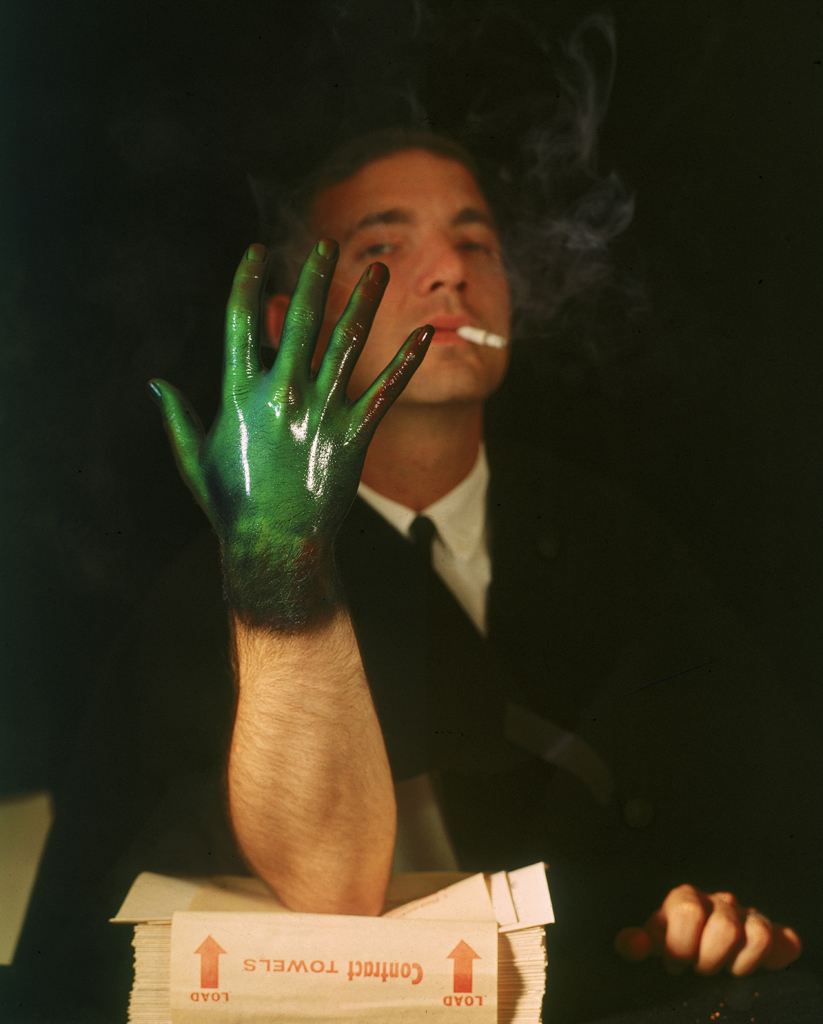 A man displays his green hand in front of his face while smoking a cigarette.