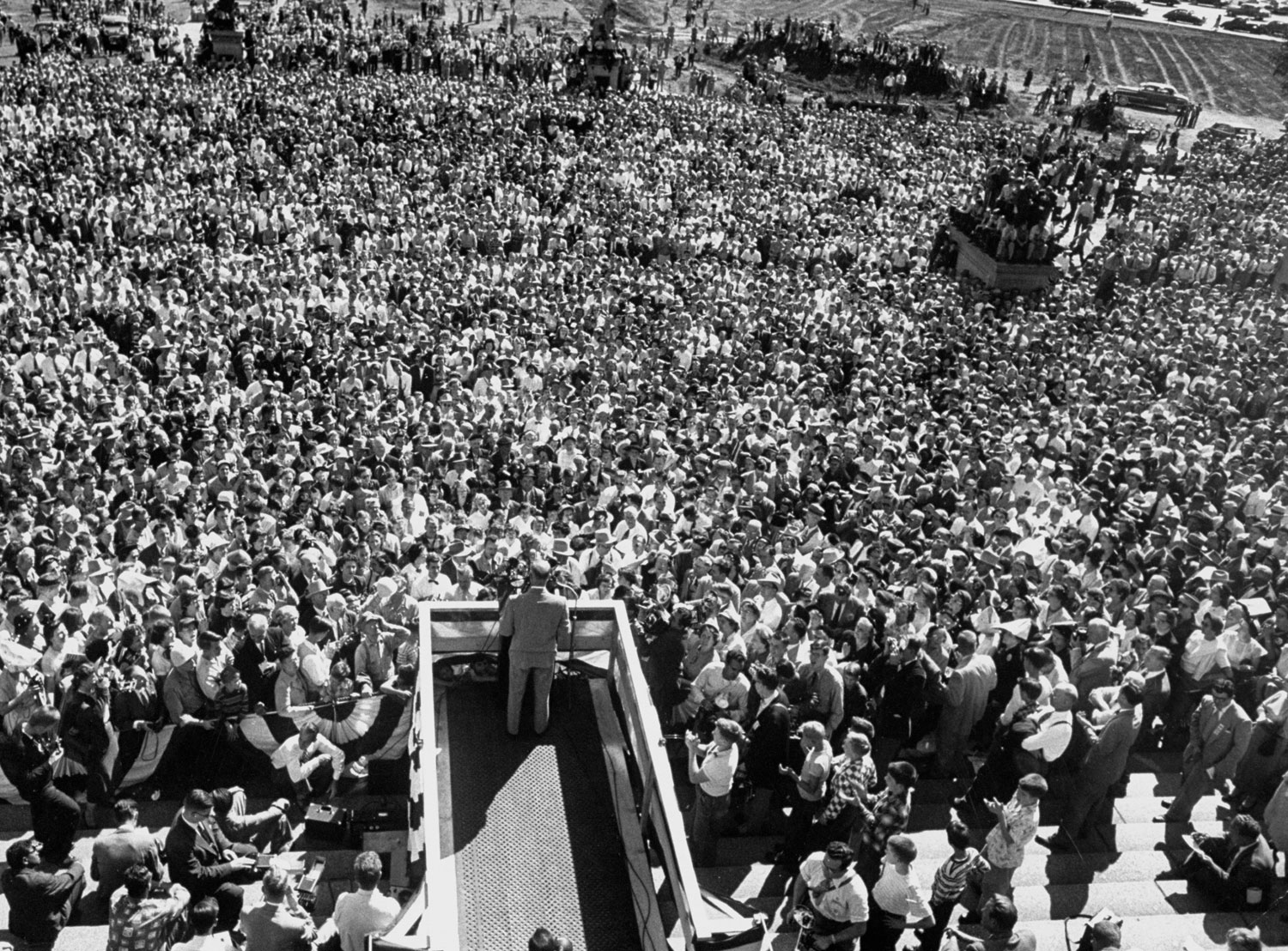A photo from above of General Dwight D. Eisenhower standing at a lectern delivering a speech during a campaigning whistle stop tour of the mid-west in September 1952. There are hundreds of people in the crowd.