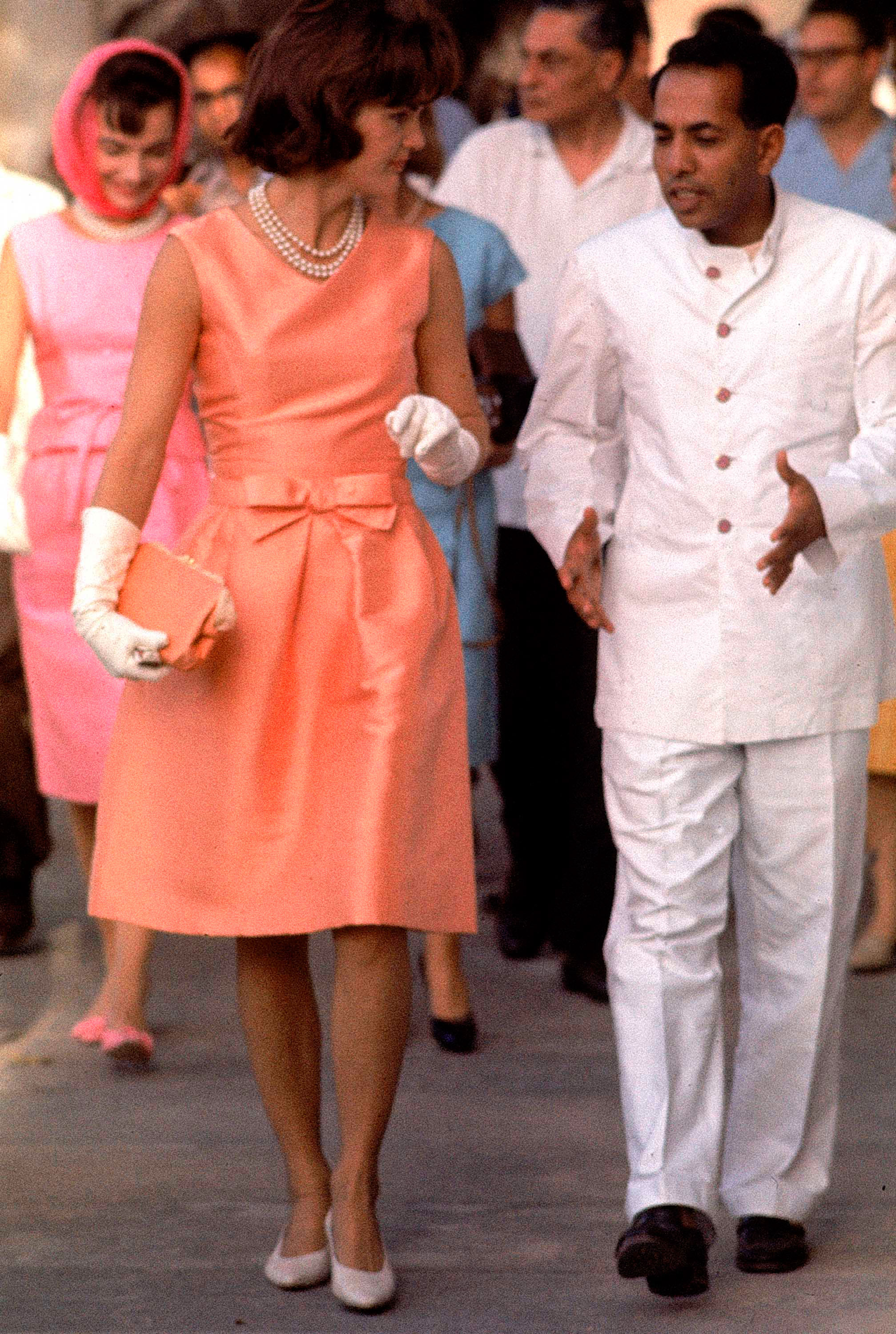 In fitted silk apricot dress, Jackie walks through crowds at Udaipur, where she was given a noisy reception." She walks with the Maharaj of Mewar, left, during her visit in 1962.