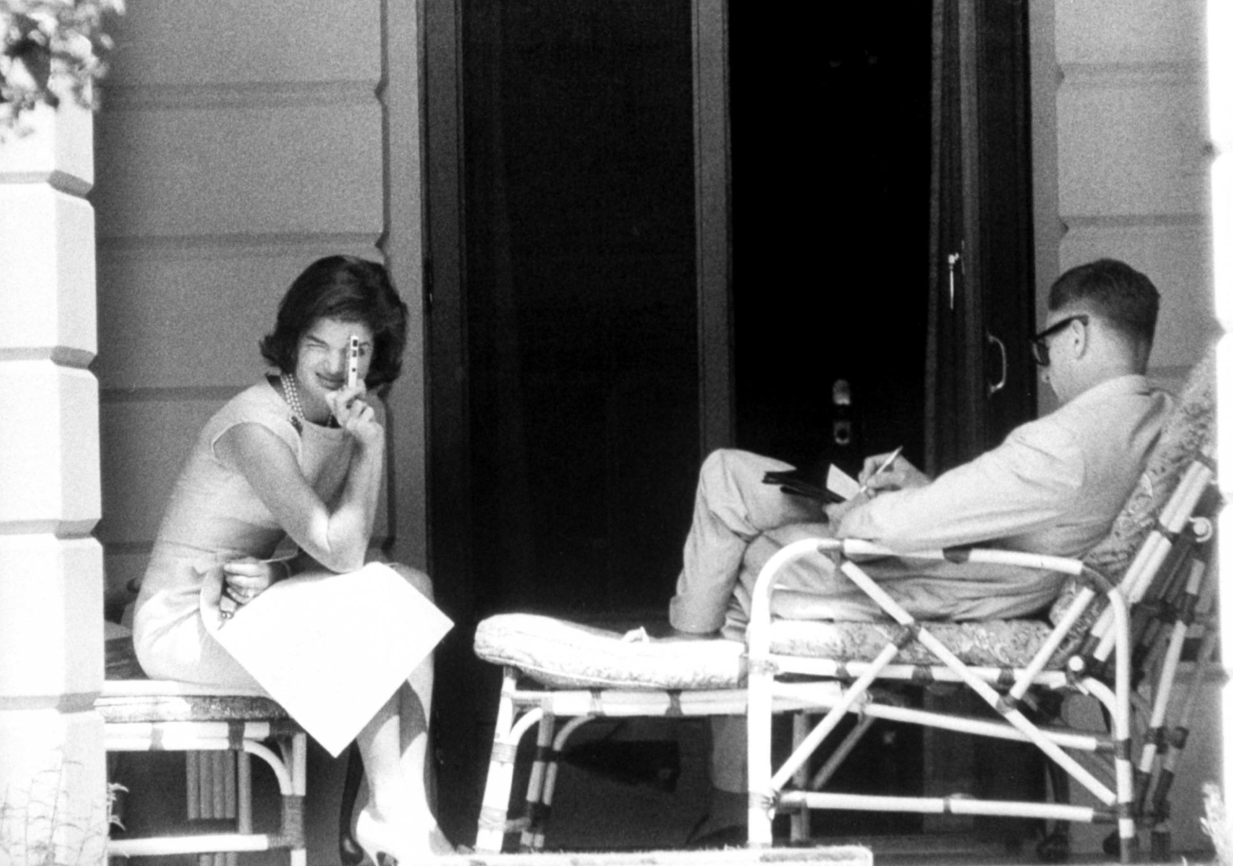 On veranda outside her room at the residence of Prime Minister Nehru, Mrs. Kennedy turns her miniature camera on photographers. Beside her, Ambassador Galbraith busies himself with his notes