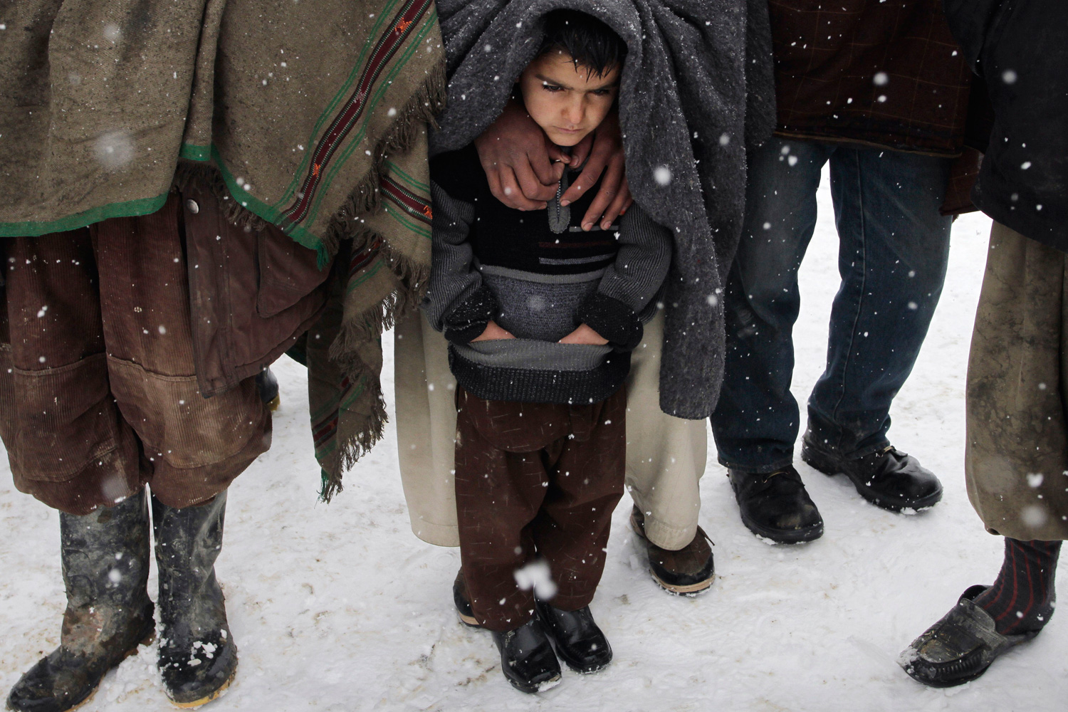 Feb. 20, 2012. A child stands with his father as they wait to receive blankets and winter jackets from Welthungerhilfe, a German NGO, during a snow fall at a camp for internally displaced Afghans in Kabul.