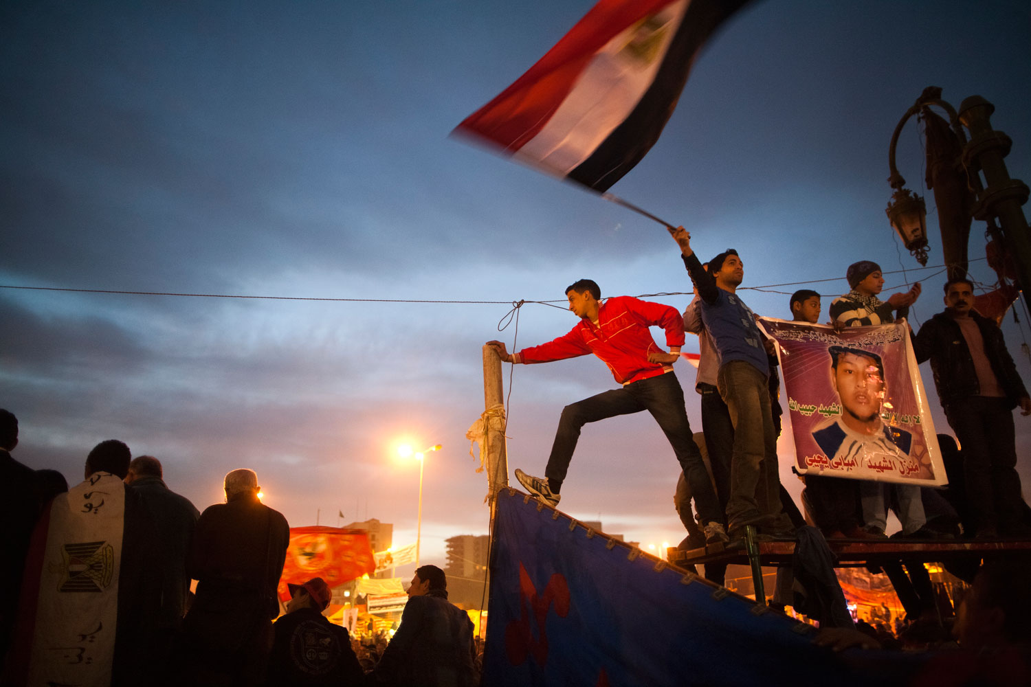 Egyptian youths watch an assembled crowd while holding a flag and a martyr poster in Tahrir Square.