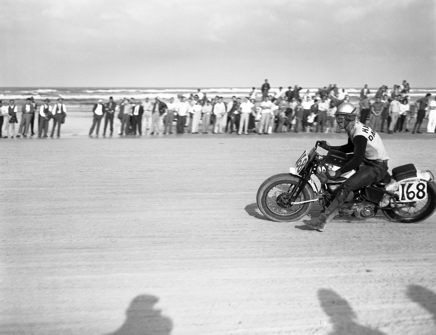 His leg outstretched for balance, a racer (number 168) steers his motorcycle across the sand during the Daytona 200 motorcycle race, Daytona Beach, Florida, March 1948.