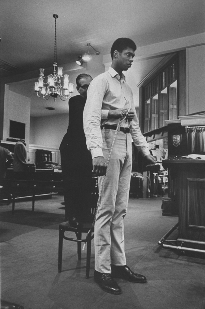 U.C.L.A. basketball star Lew Alcindor standing at 7 ft. 2 inches tall, being fitted for trousers with a 51-inch inseam by a tailor who is standing on a chair.