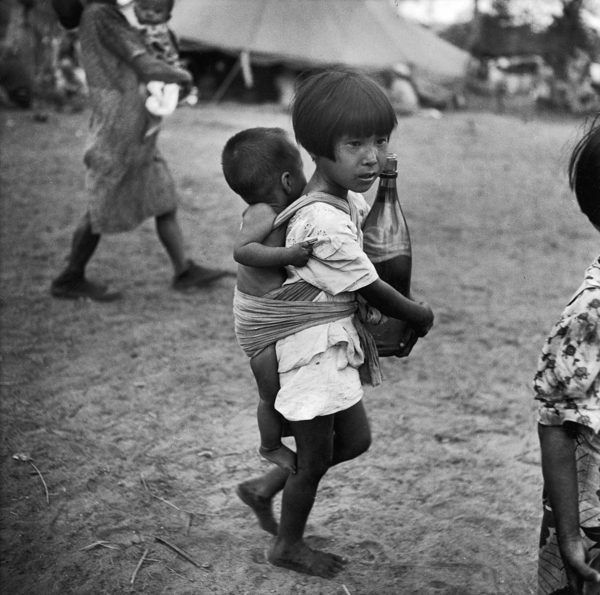 A girl in a village on Saipan (Marianas Islands) carries a bottle of water in her arms and a baby on her back in 1944.