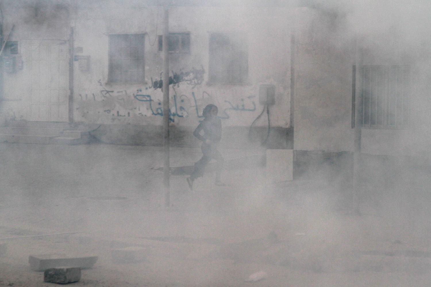 April 2, 2012. A Bahraini anti-government protester runs from tear gas, past a wall covered in anti-government graffiti, during clashes with riot police in Salmabad, Bahrain.