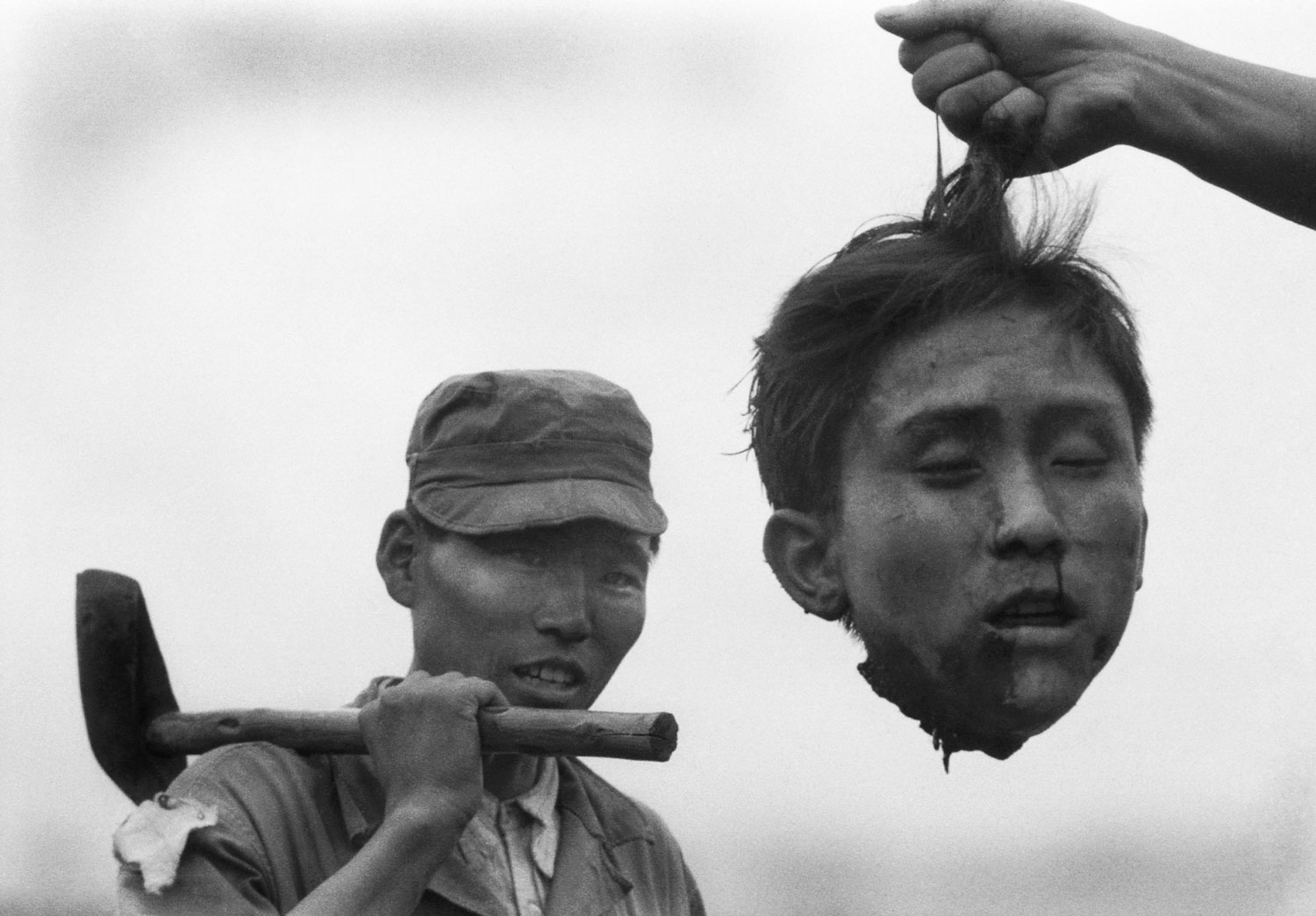 Hand of unseen South Korean holding severed head of North Korean Communist guerrilla by his hair as a member of the South Korean National Police, smiles broadly, w. an axe over his shoulder.