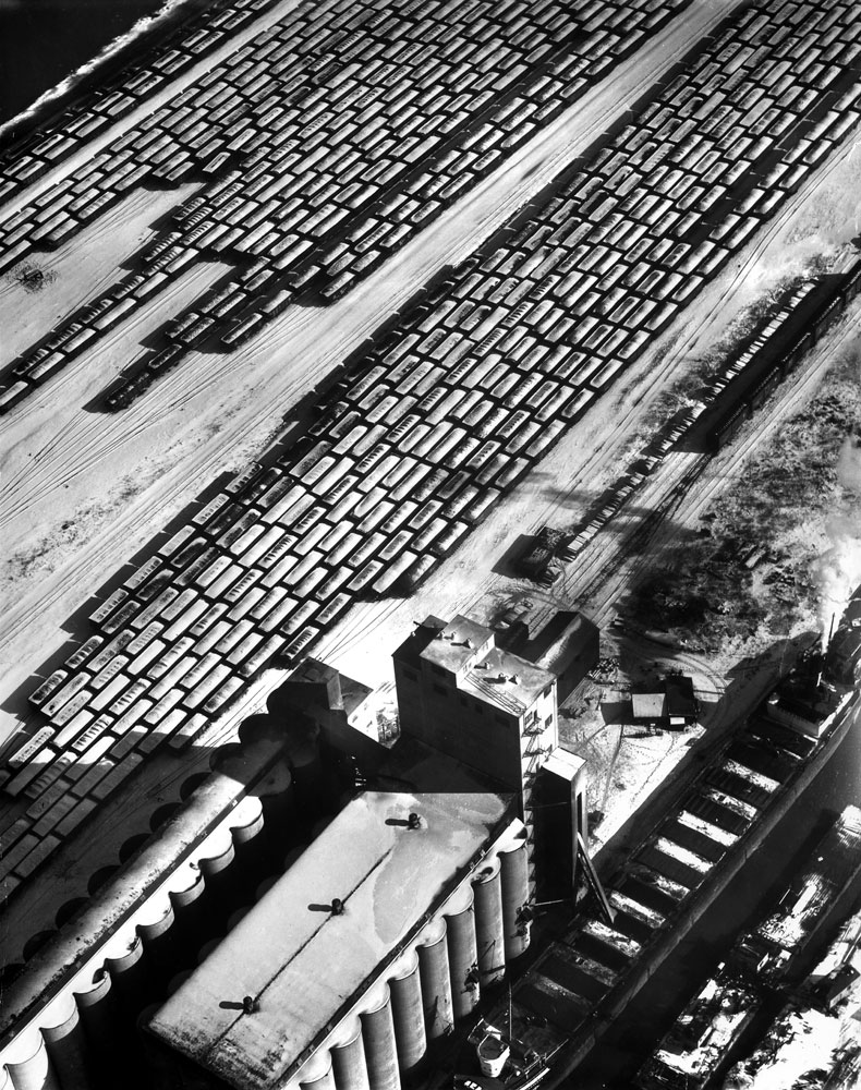Grain elevator, operated by the Norris Grain Co. on the southeast side of Chicago, unloads corn from lake boat in a Calumet River slip (right foreground). In the freight yards (background) snow-covered gondola cars are loaded with coal.