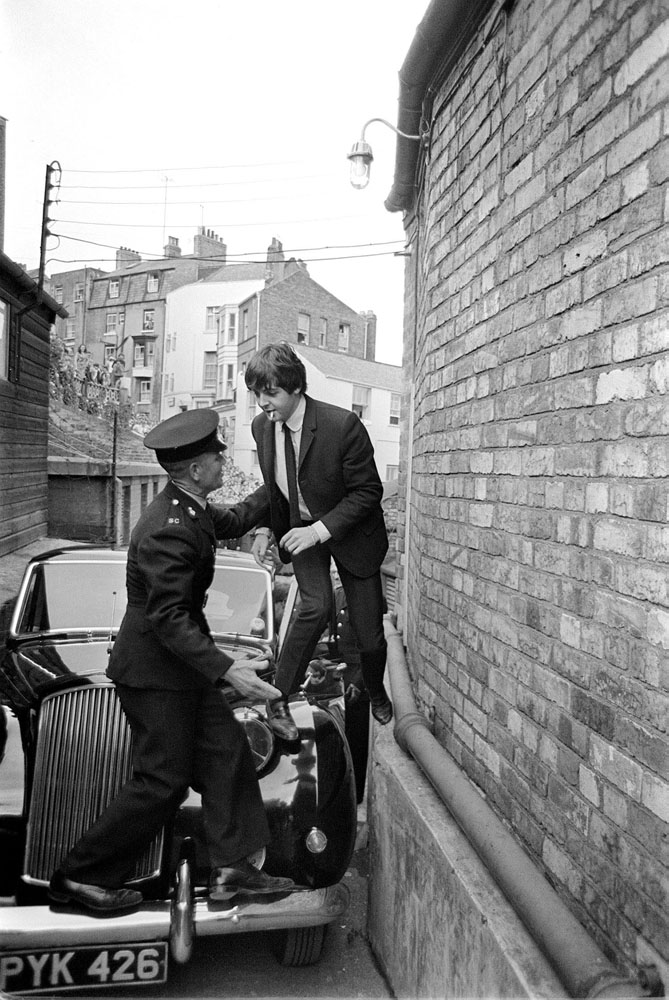 Escaping fans Paul McCartney is helped over his car by a guard before The Beatles concert at the Futurist Theatre. Fans can be seen in the background, Scarborough, England, 1964.