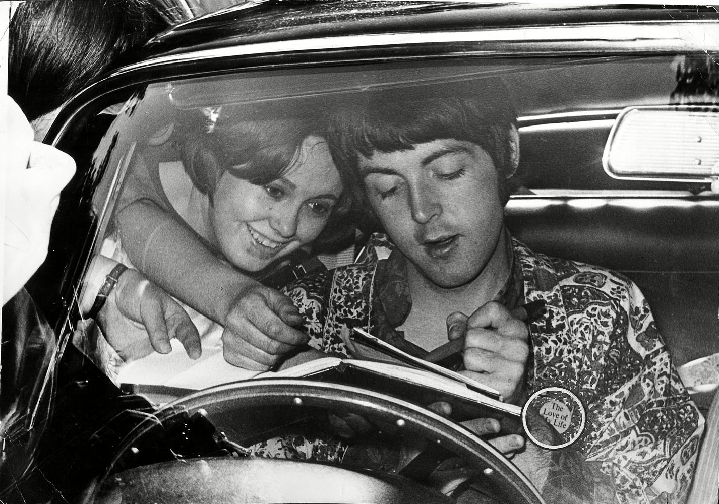 Paul McCartney signs autographs for fans whilst sat in his Aston Martin car outside his flat in St John's Wood. He has a badge on his shirt which reads 'The Love Of My Life'. London, England, 1967. (In this photo, the circle highlighting the badge was made by a prior media source)