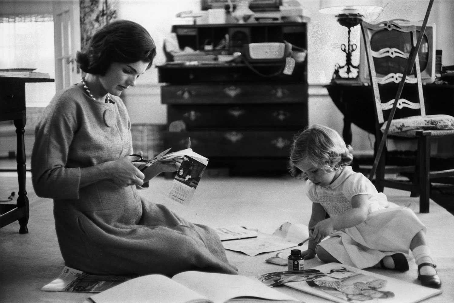 Jackie Kennedy cuts newspaper clippings next to an open scrapbook as her daughter Caroline toys with the applicator from a glue bottle, 1960.