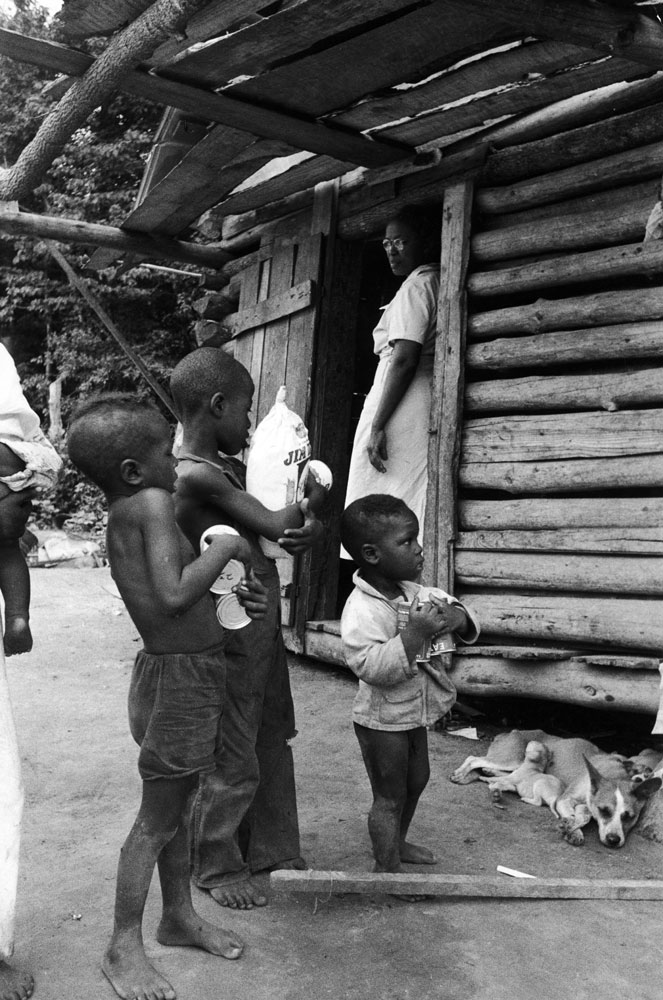 Caption from LIFE. Store-bought-food donated by Maude fascinates youngsters outside log cabin. ...