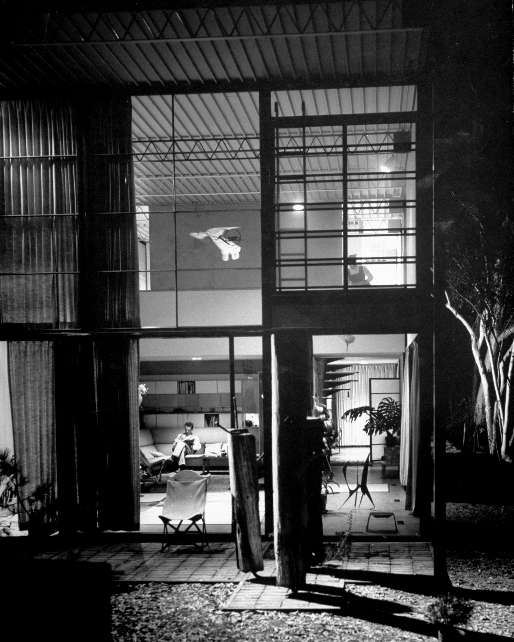 Interior view shows living room's 17-foot-high ceiling, unadorned steel-truss construction, to which Eames clamps lamps for varied lighting effects. He puts up the pilings from an old pier outside the door because he liked their looks. He similarly suspended a Chinese owl kite and toy French horn from the ceiling.