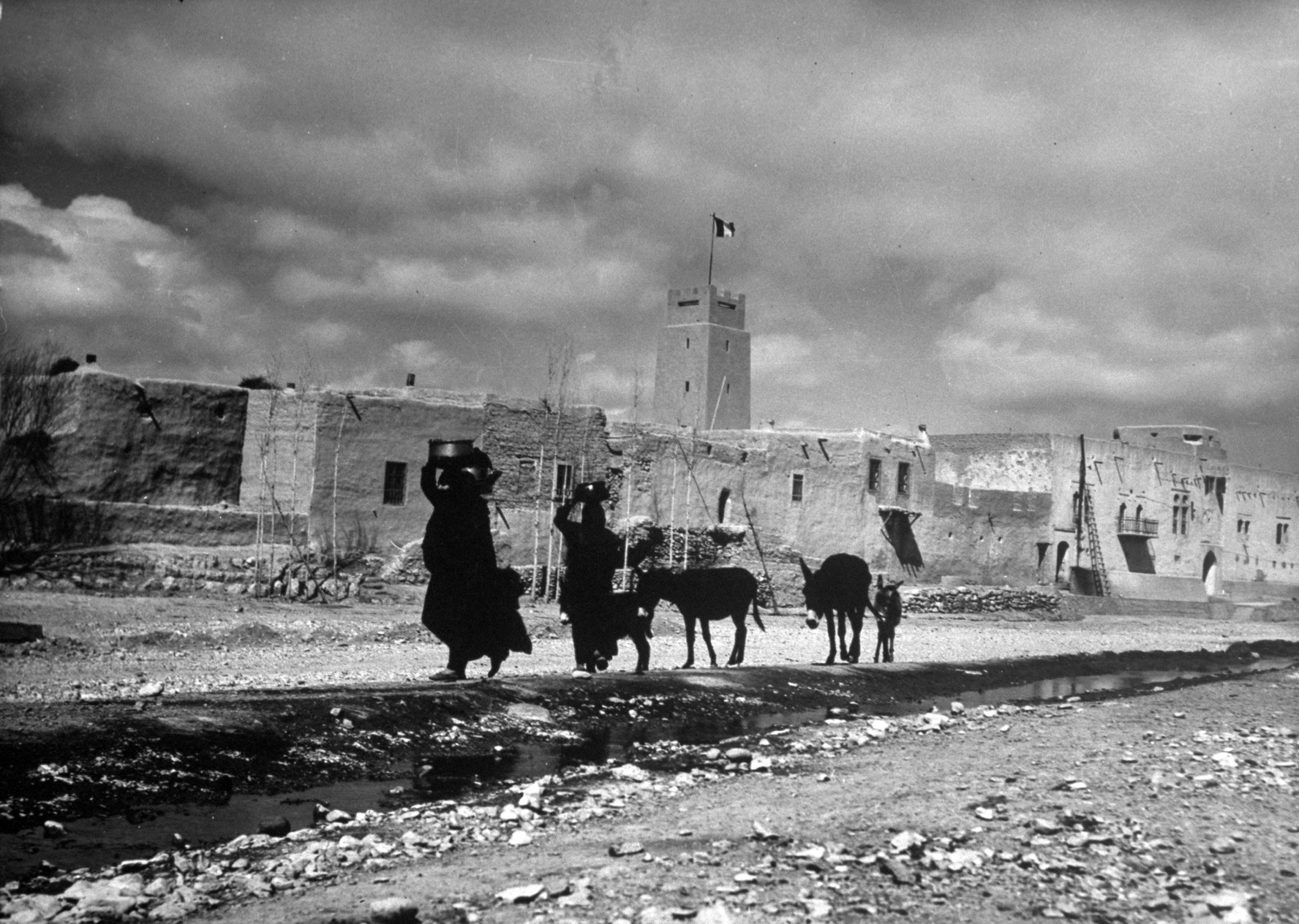 Women carry water containers on their heads as they lead mules along a road in a desert village near Damascus, Syria, 1940.