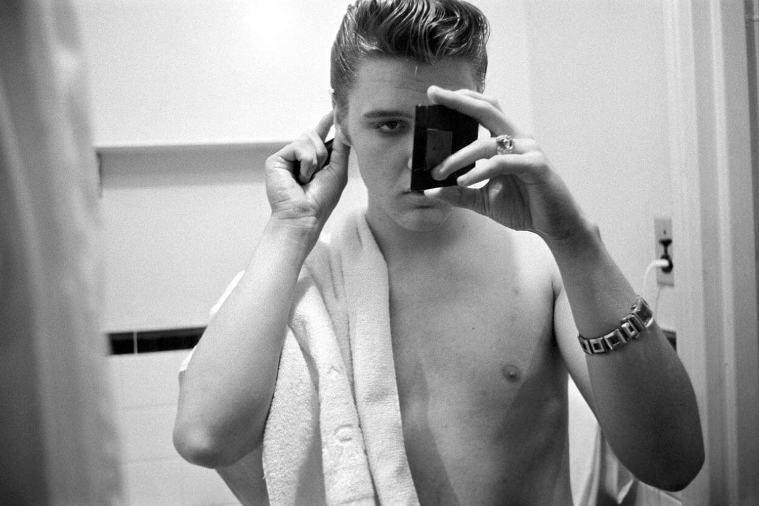 March 17, 1956. The Warwick New York. Elvis in his hotel bathroom an hour before returning to perform on 'Stage Show.' I love this one…he’s got full concentration. Elvis had just finished showering — he's looking into a small ladies hand mirror to see what's going on with the back of his hair, in a bigger mirror. He's using Vaseline hair tonic, not  bear grease.