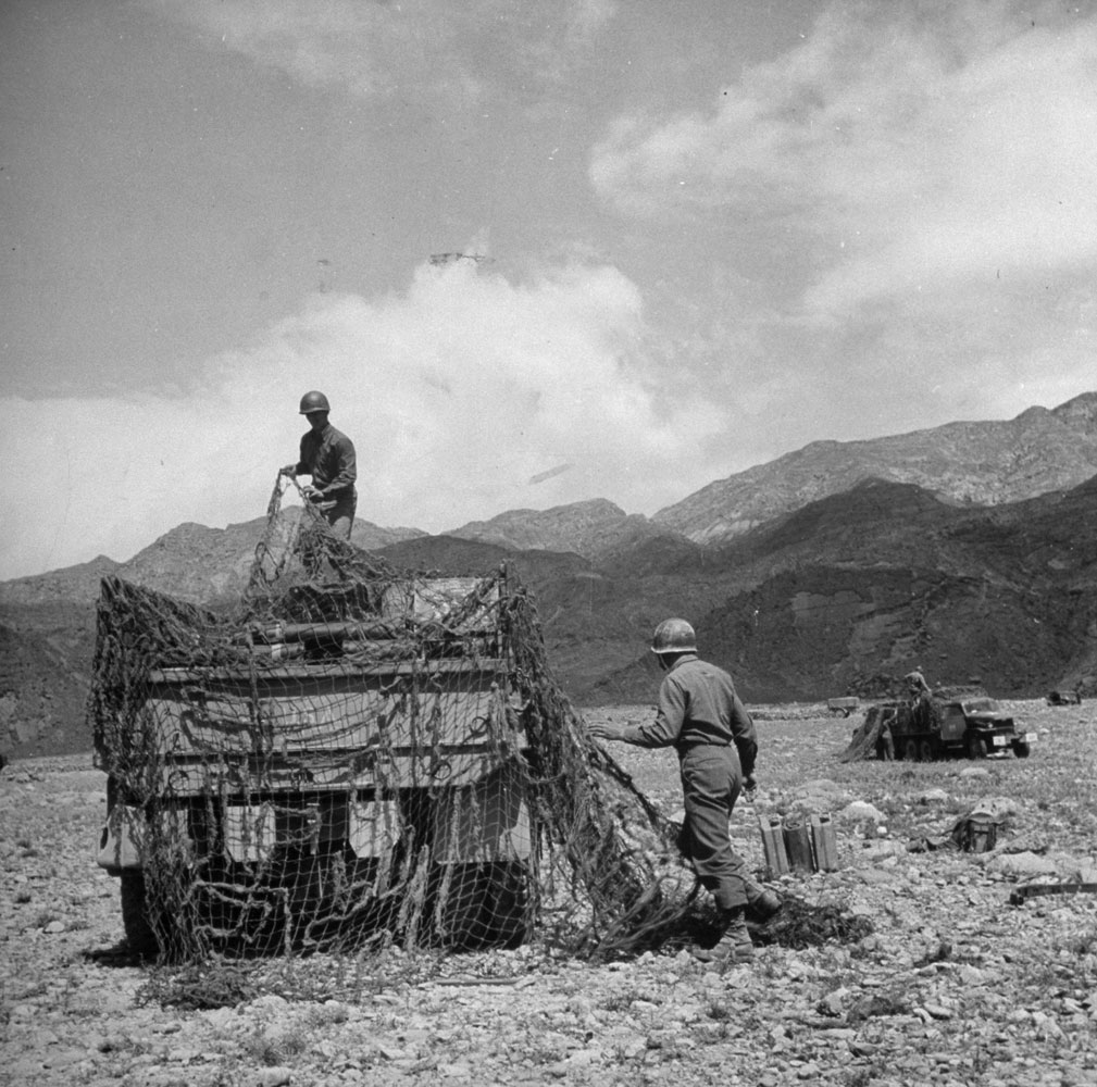 Covering two and a half ton truck with net after arrival at ammo dump near front. Ammo is moved to artillery at night. Ammo is 105mm in clover leaf (cluster of 3 shells).