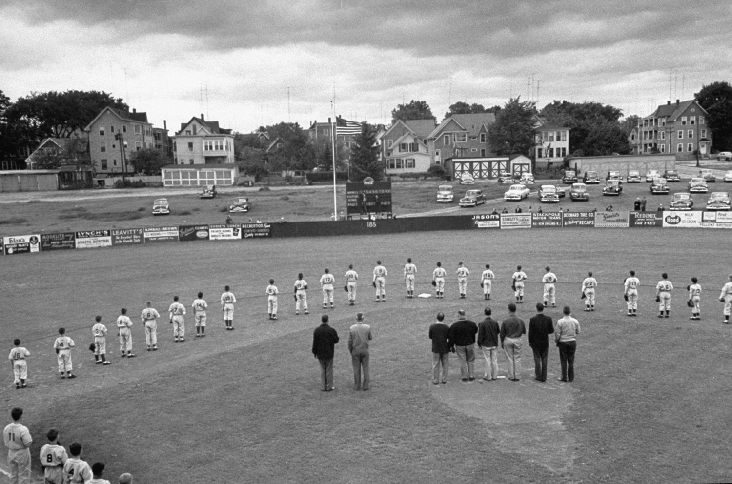 Before game the players and coaches stand at attention and face flag during national anthem. Little League field is patterned after major league stadium but in almost every detail is one third smaller.