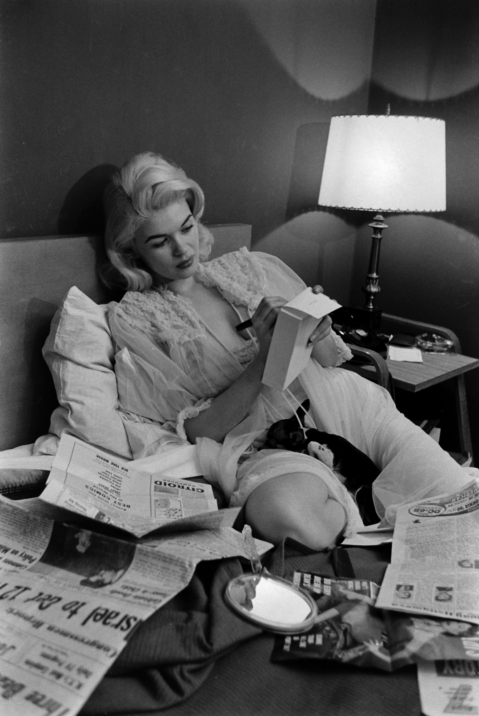 Jayne Mansfield, clad in a revealing negligee, looks over newspapers with her pet Chihuahua in her lap, Hollywood, 1956.