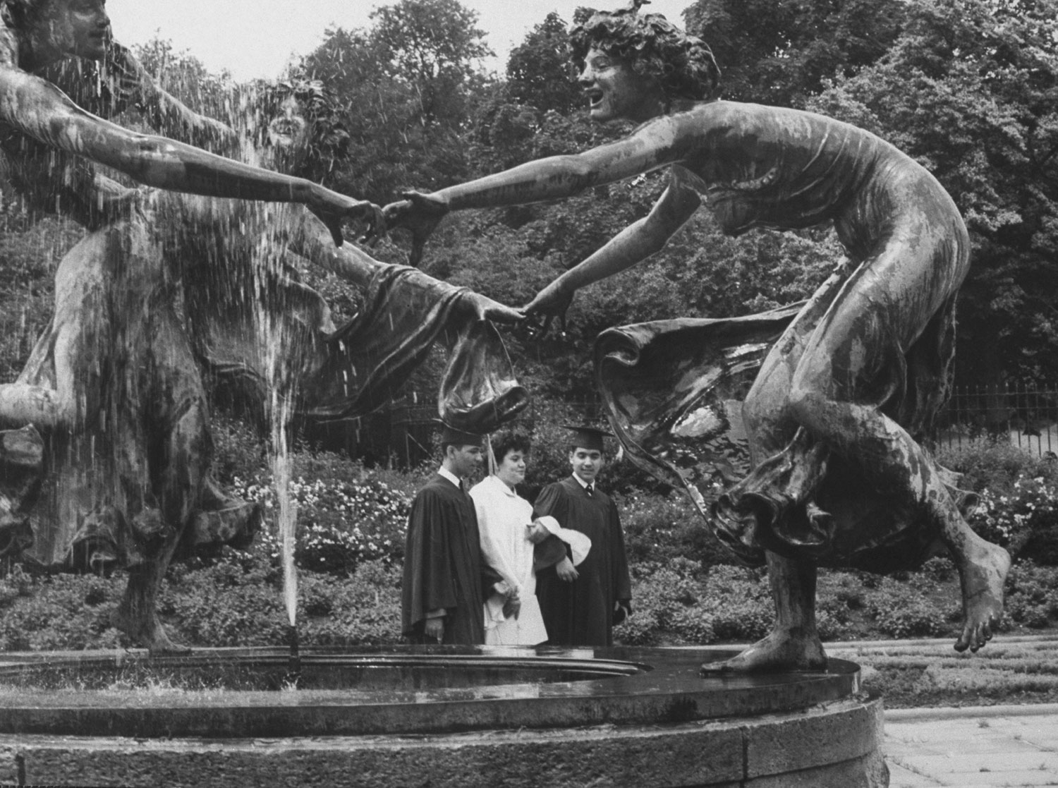 A trio of newly graduated students strolls soberly past a trio of figures dancing ring-around-a-rosy at a fountain.