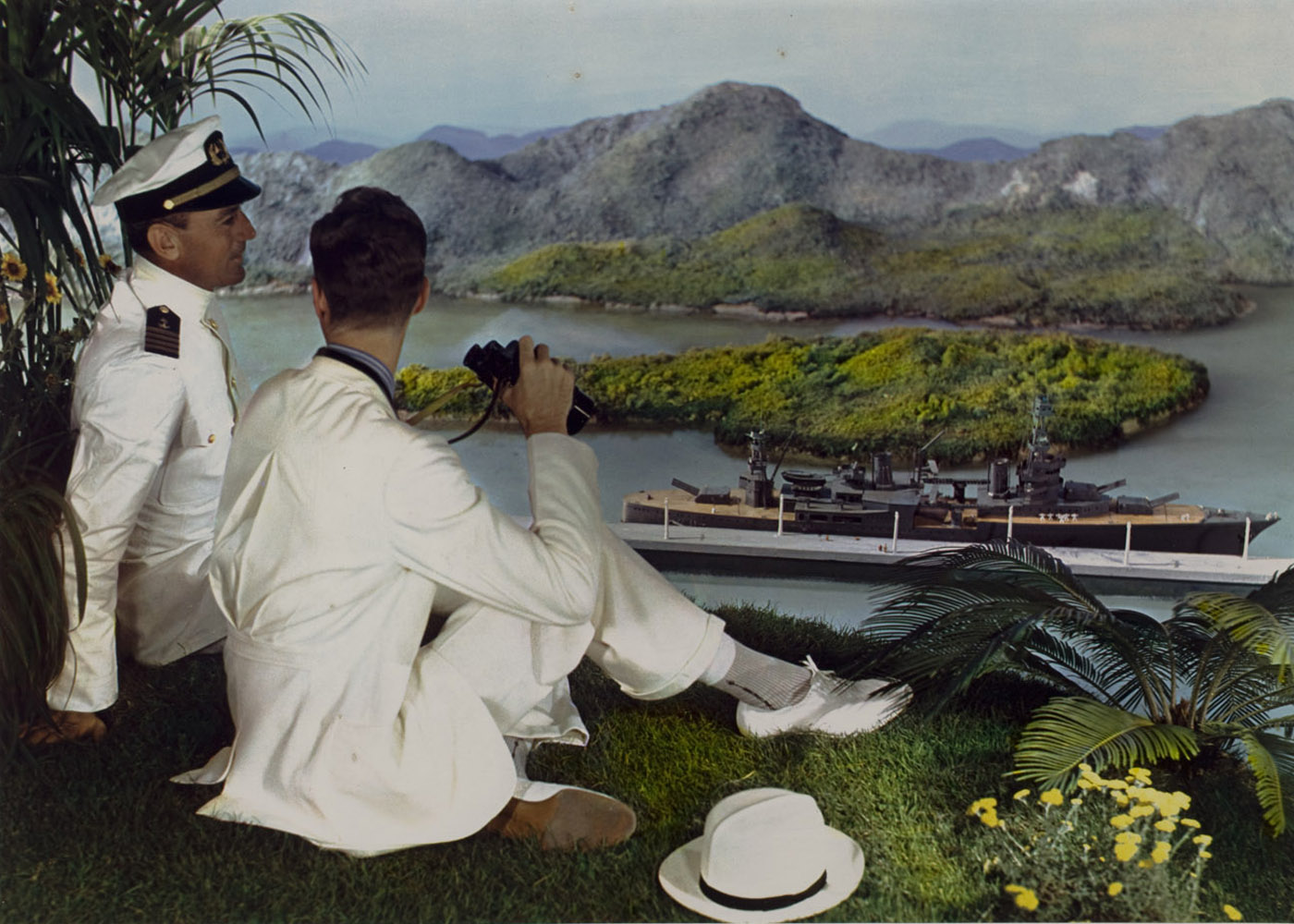Naval officer and young man on hillside with binoculars, overlooking battleship in bay below, ca. 1950