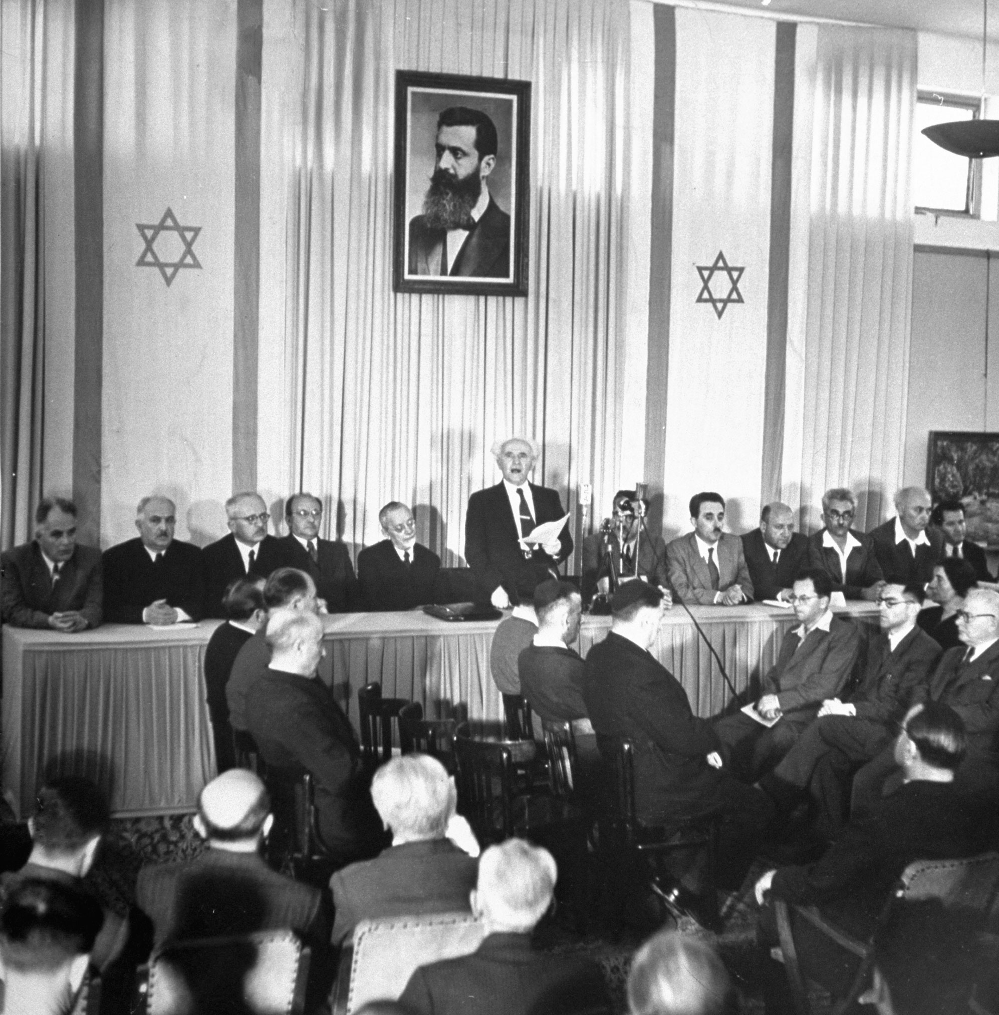 Proclamation of Nationhood is read by Israel's Prime Minister David Ben-Gurion. Around him are members of the provisional government, including Foreign Minister Moshe Shertok (third from right). Labor Minister Moshe Ben Tov (extreme right) wears sport shirt. Portrait above is of Theodor Herzl, Zionism's founder