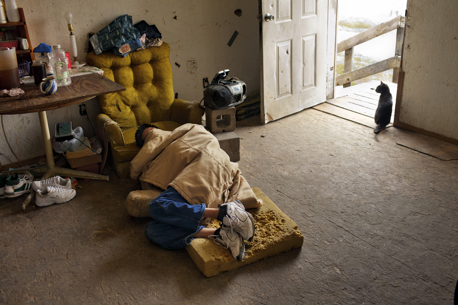 A young man suffering from the effects of a neurological disease and alcoholism sleeps in the living room of his home, six miles from the nearest town. Since the photograph was made, in May 2011, the house has been condemned, and he and the other occupants have moved elsewhere.