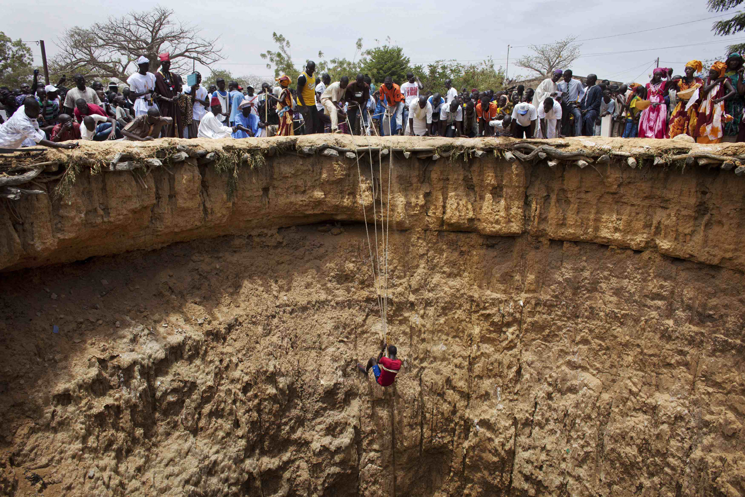 People watch a man descend into a large former well during a traditional ceremony in the village of Ndande