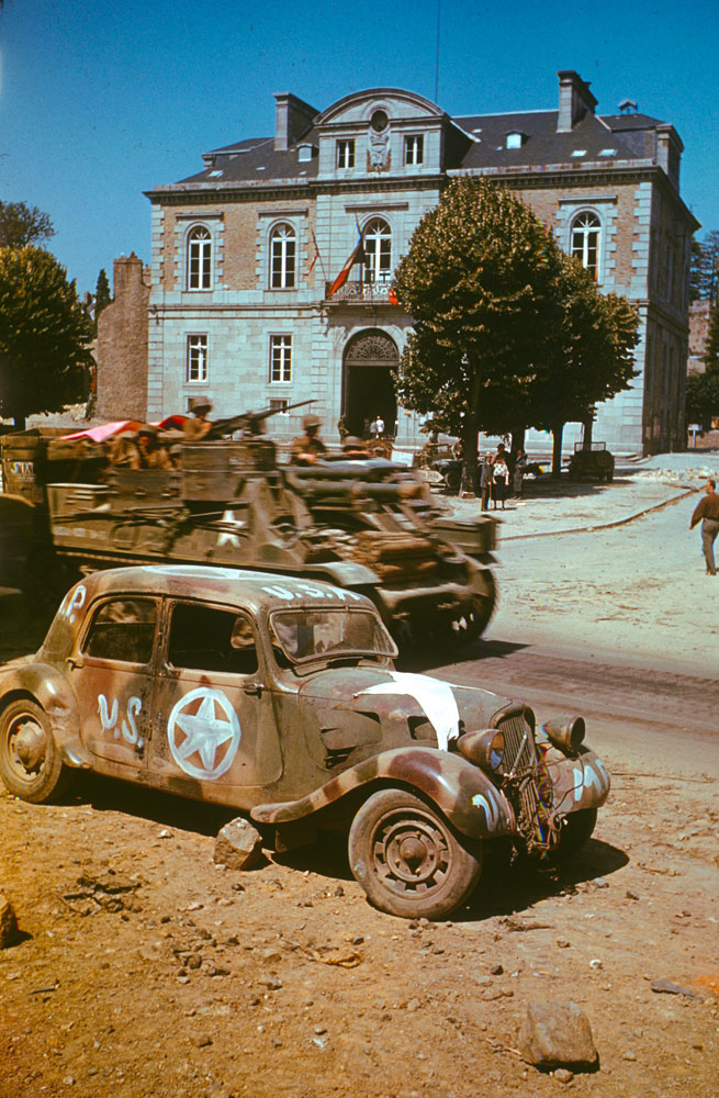 Armored vehicles on the move past civic buildings in Avranches, 1944.