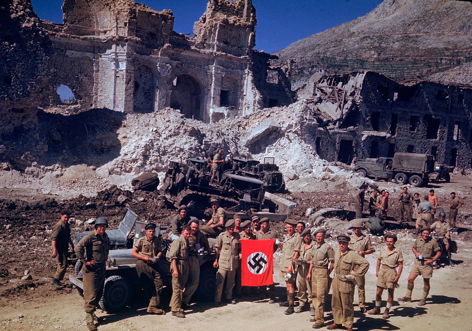 British and South African soldiers hold up a Nazi trophy flag while combat engineers on bulldozers clear a path through the debris of a bombed-out city, Italian Campaign, World War II.