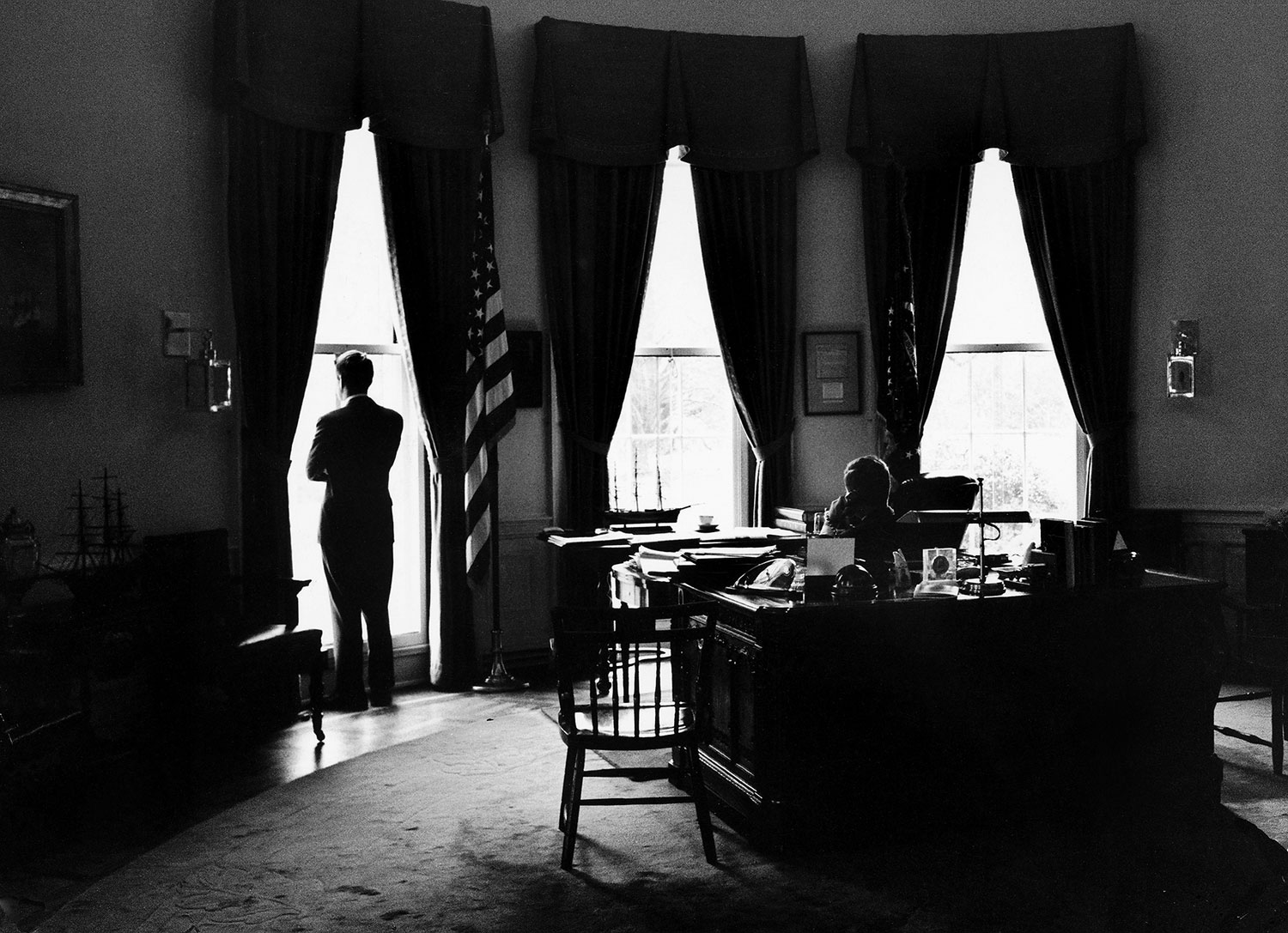 President John Kennedy and Attorney General Robert Kennedy (standing at window) in the Oval Office, 1962.