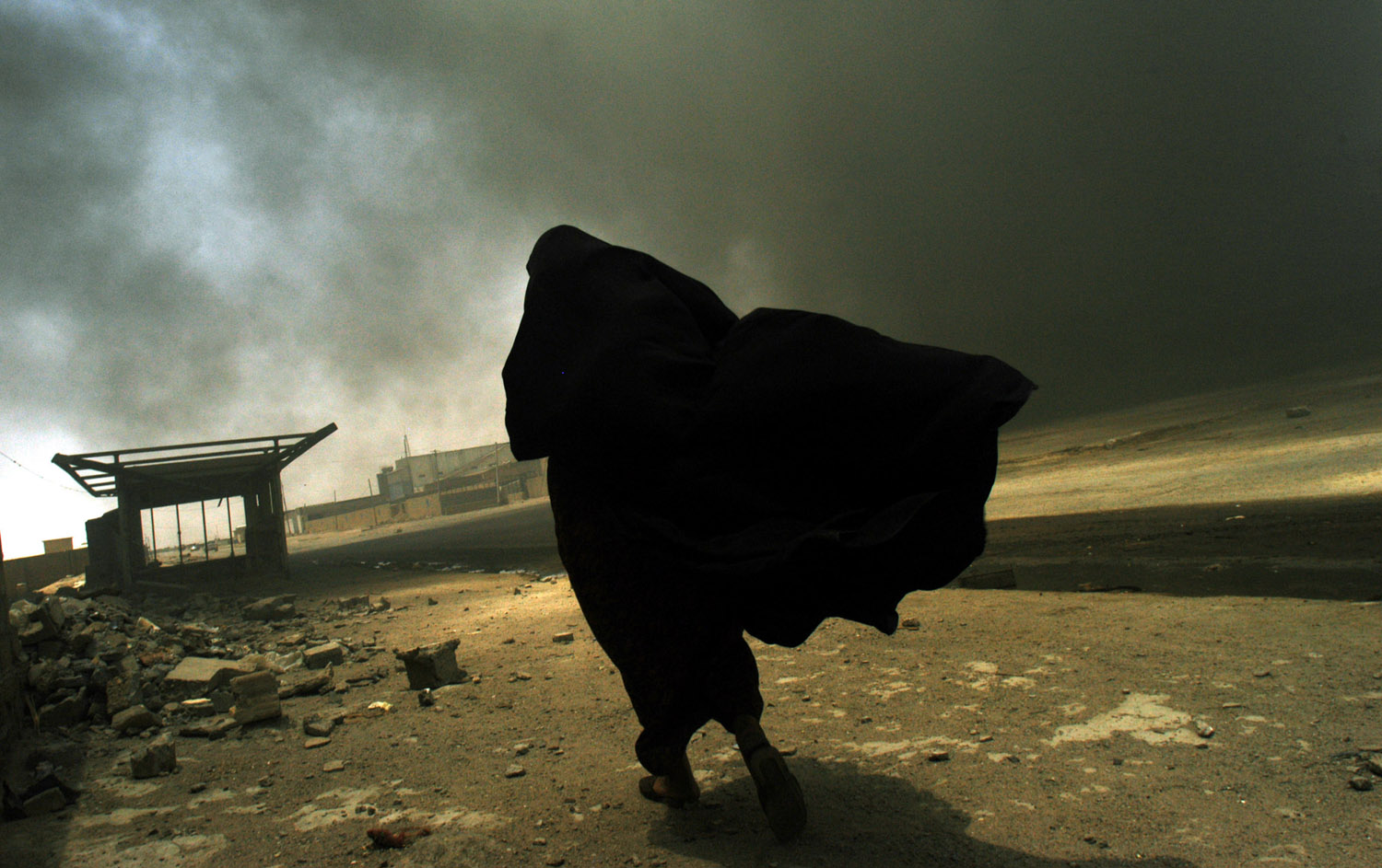 An Iraqi woman walks through a plume of smoke rising from a massive fire at a liquid gas factory as she searches for her husband in the vicinity of the fire in Basra, Iraq, May 26, 2003.  The fire was allegedly started by looters picking through the factory, and residents in the vicinity feared the explosion of the four liquid gas tanks on the premisis.   Weeks after the end of the war, looting continues to be one of the main problems for both Basra and Bagdad cities as coalition forces struggle to get life back to normal.  (Credit: Lynsey Addario/ Corbis Saba)