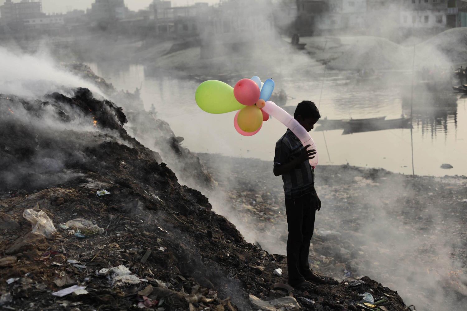 A boy plays with balloons by Buriganga river as smoke emits from a dump yard during sunset in Dhaka
