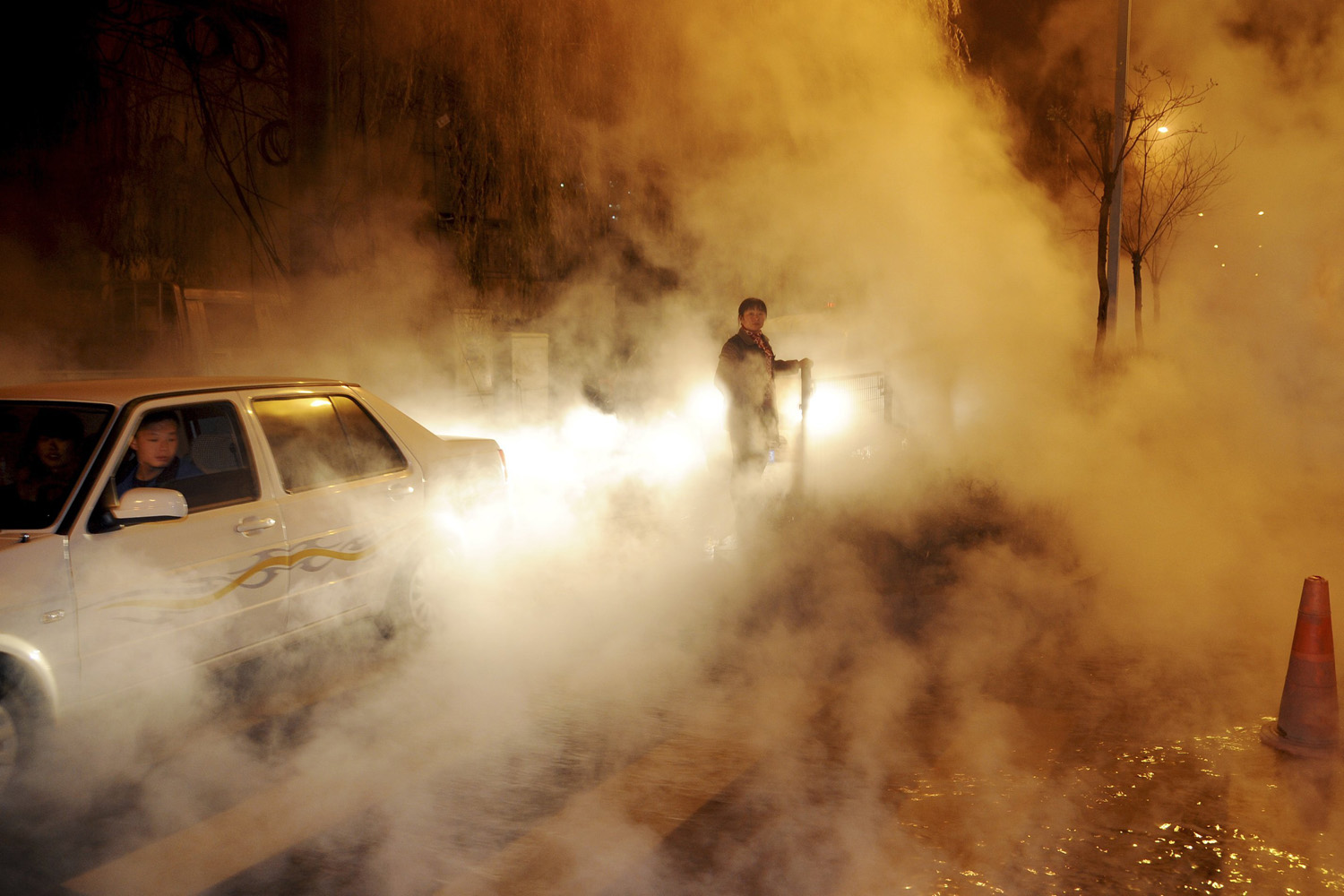 A car drives past as a woman standing amidst thick steam after hot water leaked from a heating pipeline onto a street in Taiyuan