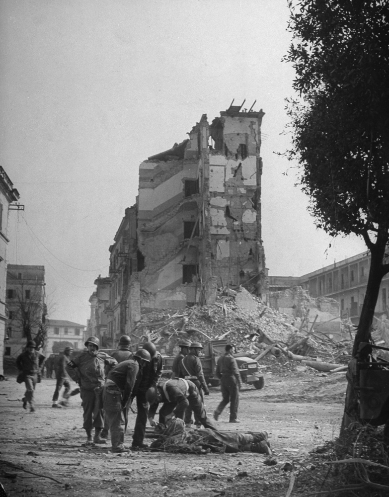 In ruined Anzio American and British soldiers gather around man who has just been hit by fragments of a shell bursting in the street. Casualty had come ashore from the harbor 40 seconds before.