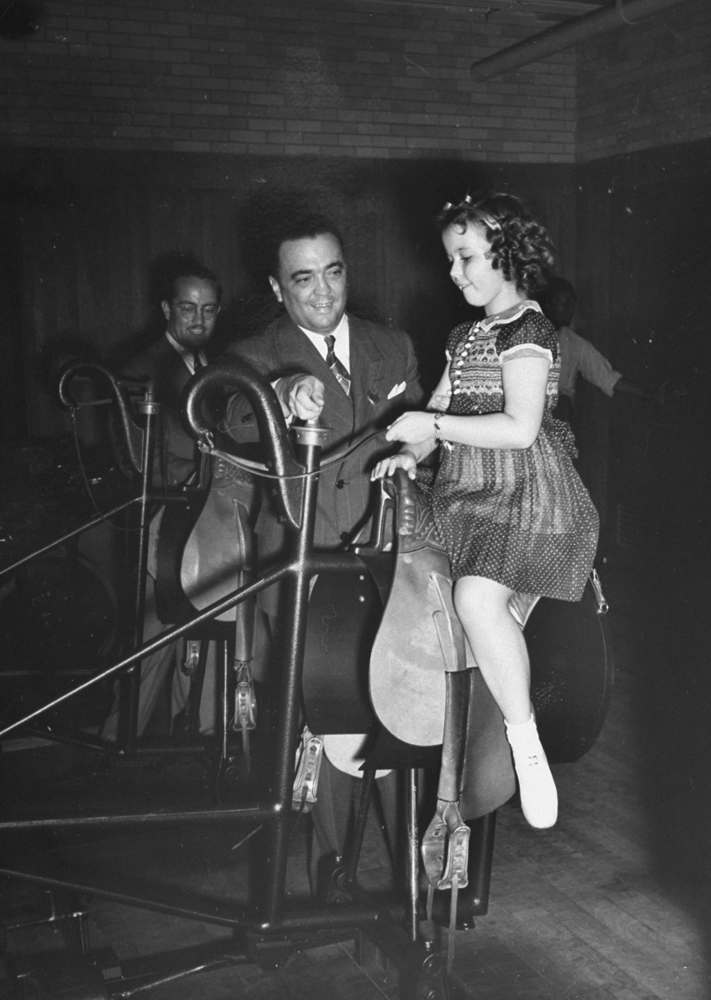 Federal Bureau of Investigation director J. Edgar Hoover shows Shirley Temple how to ride a mechanical horse, 1938.